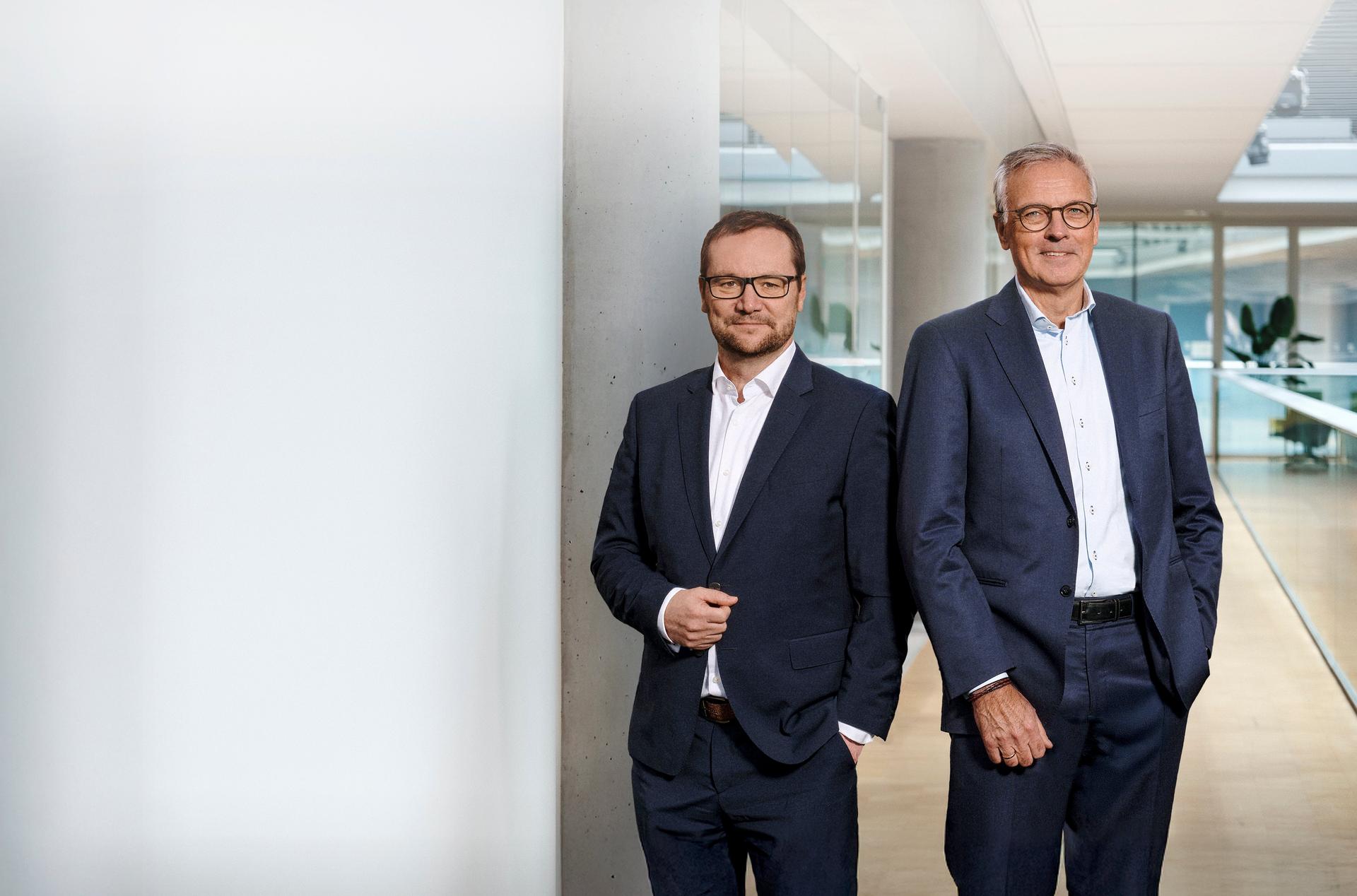 Jens and Claus, are standing in a modern, well-lit office environment. Both are wearing dark blue suits with white shirts, conveying a professional, corporate atmosphere. The background features glass windows and doors, reflecting light and showcasing a few plants.